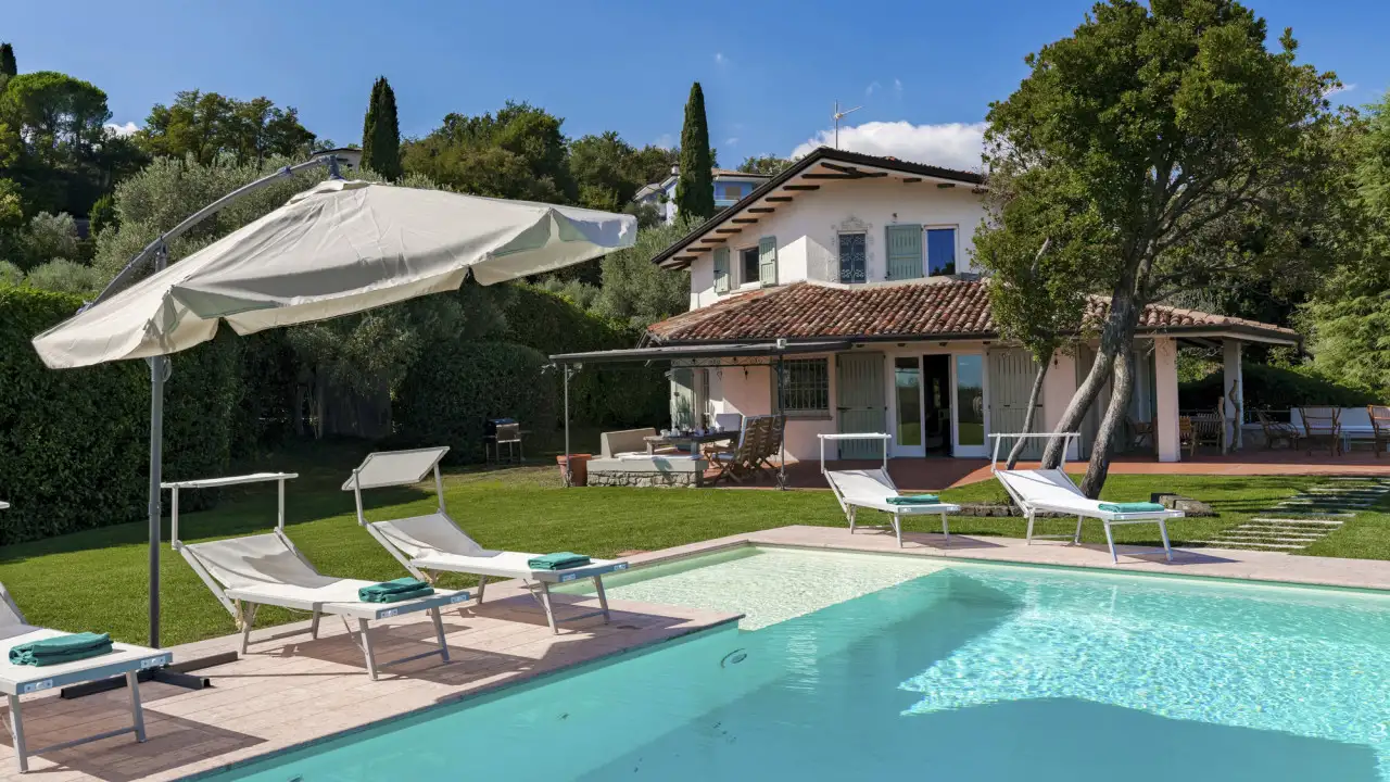 Schwimmbad mit Liegestühlen und Sonnenschirm vor Villa mit Garten und Bäumen
